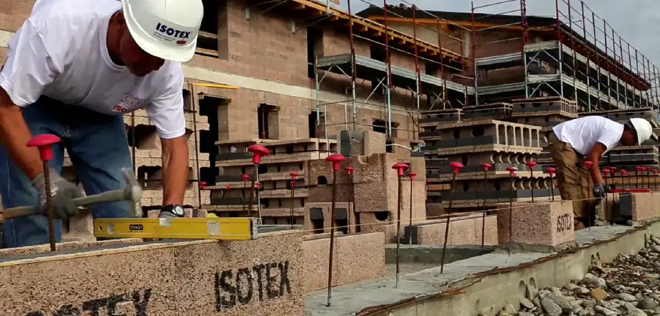 Isotex installers laying the first course of insulated wood-cement blocks with a spirit level and exposed reinforcement, a multi-storey Isotex building under construction behind.