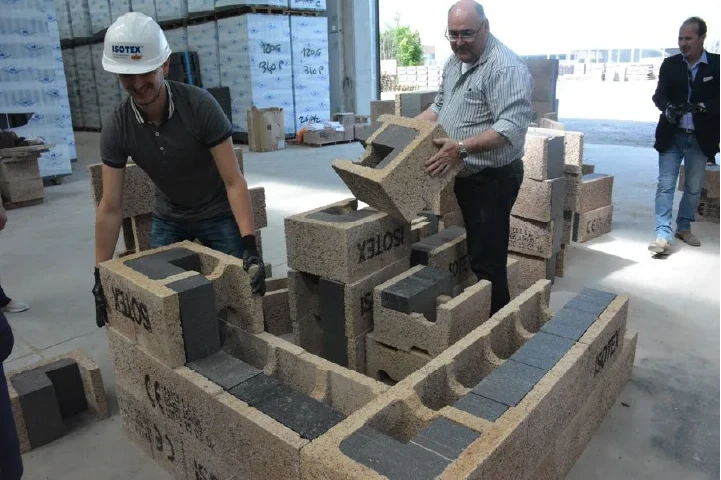 Isotex training: an installer assembles a demonstration wall of interlocking insulated wood-cement blocks while a trainer guides him at an Isotex facility.