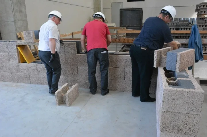 Isotex training: a small team lays a course of insulated wood-cement blocks along a wall, with reinforcement and a floor beam in place.