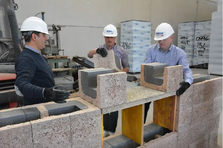 Isotex training: three people examine an insulated wood-cement block at a workbench, the U-shaped channel block and core visible.
